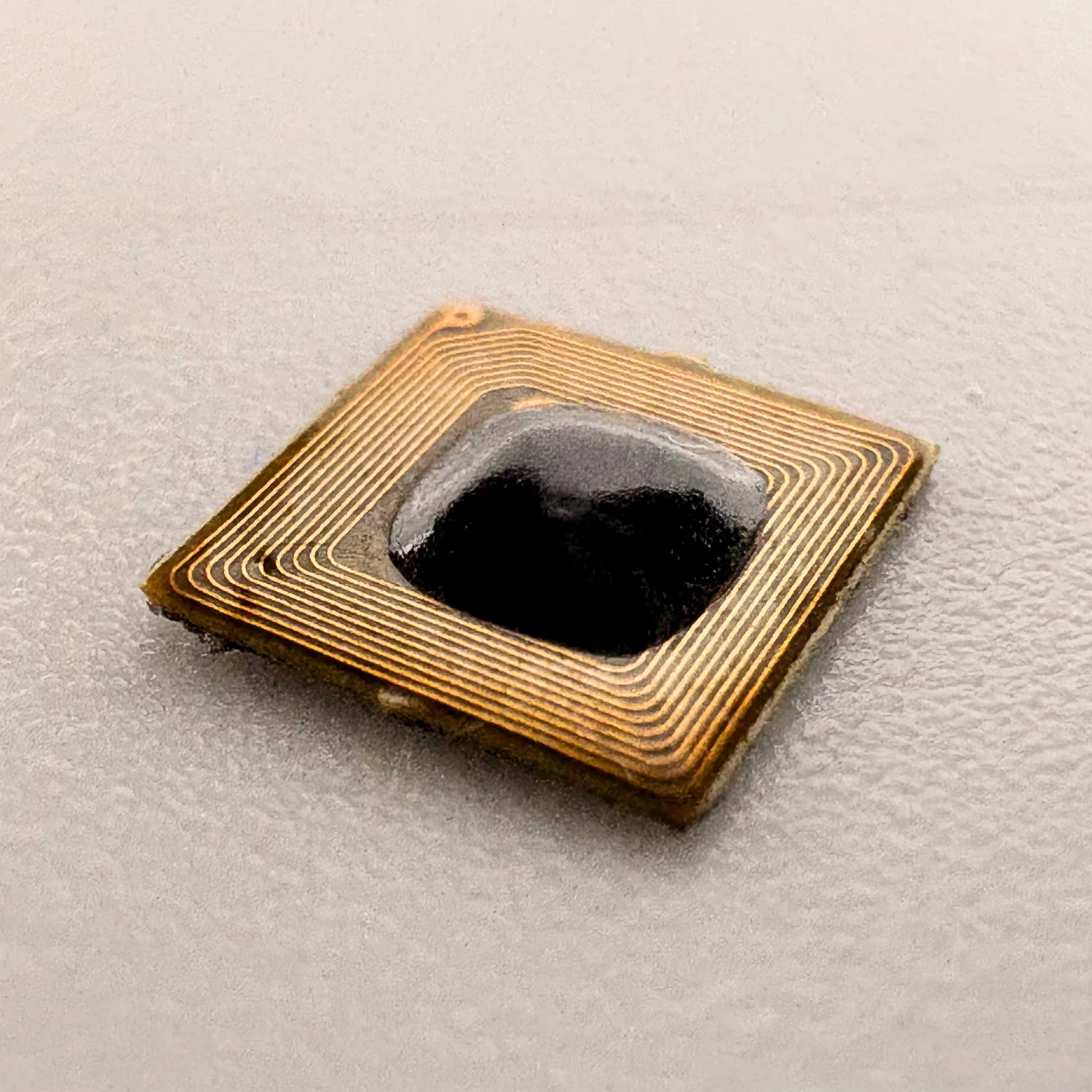 Close-up of a small, square NFC chip with gold-colored circuit lines on a gray background.